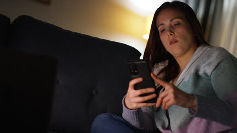 Mujer-Sentada-En-Un-Sofá-En-Casa-Por-La-Noche-Transmitiendo-O-Viendo-Una-Película-O-Programa-En-Una-Computadora-Portátil-Y-Navegando-Por-Internet-En-El-Teléfono-13